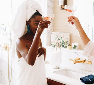 A model wears a white bath towel around her hair and another around her body while she cheers with a glass of rose wine.