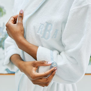 A model wears a white bath robe with light blue piping and  monogram. She buttons the cuff of her sleeve.