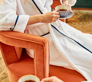 A model wearing a white robe with navy piping sits in an arm char and holds a cup of tea,